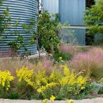 A garden with yellow flowers and pink grasses bursts with blooms in front of a grey metal seed silo