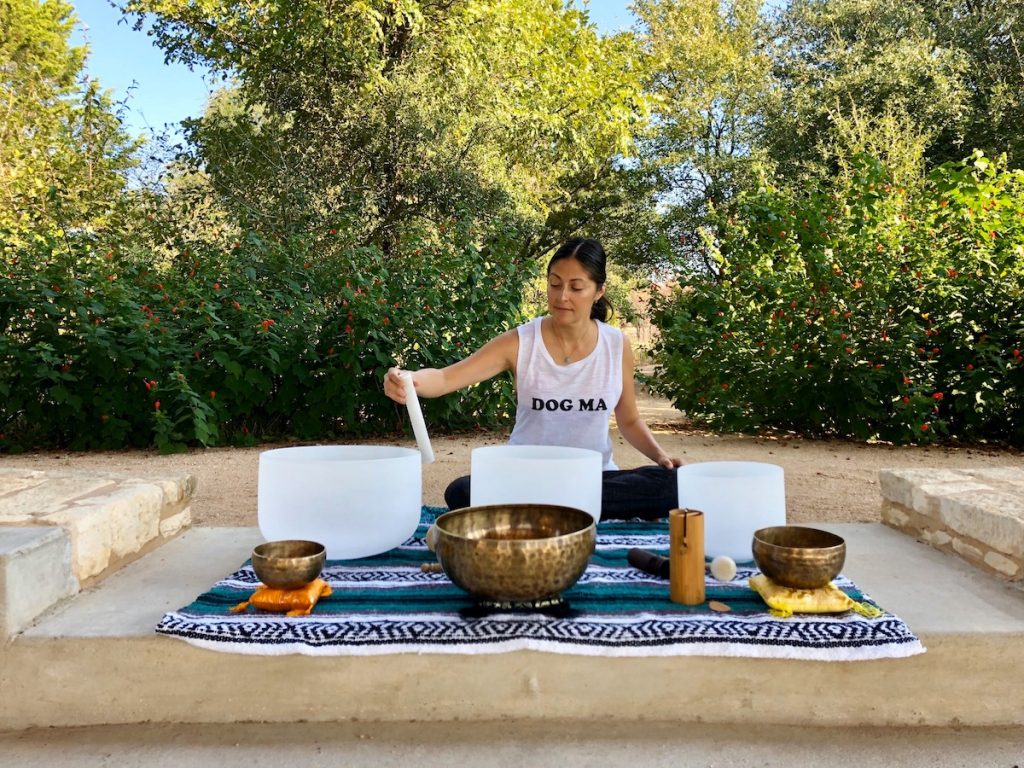 A yoga instructor, Cynthia Bernard, wearing a "Dog Ma" white tank and stirring the singing bowls laid out on a blue blanket in the gardens for a yoga class.