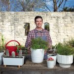 Smiling man standing behind an array of plants in white pots and a red watering can