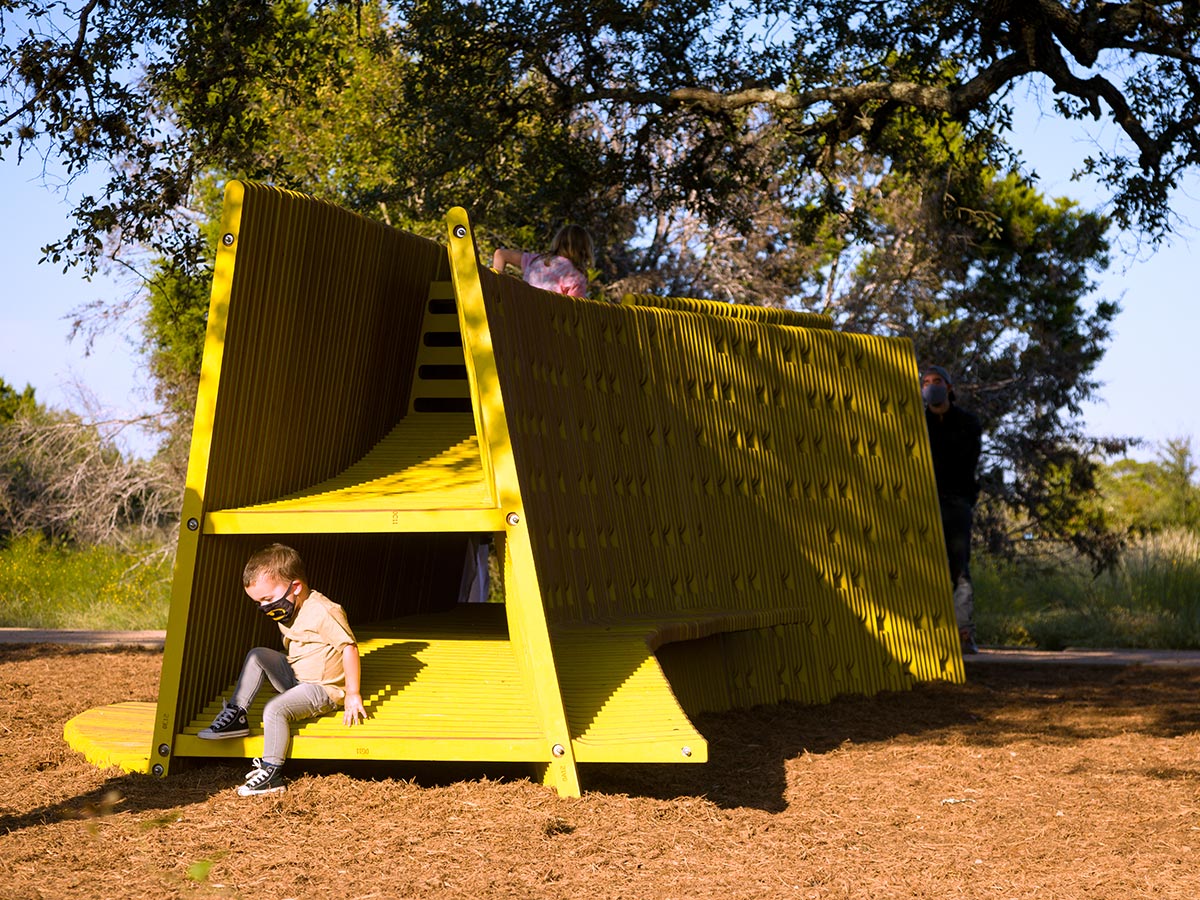 A small child wearing a face covering sits inside a yellow A-Frame structure atop a bed of mulch and under a large oak tree.