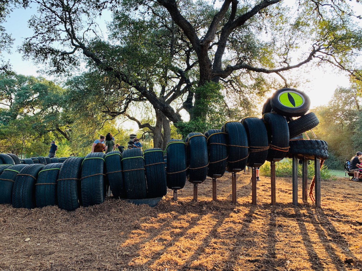 A structure or sculpture of a large snake made of car tires with a bright green eye and rope tongue with mulch on the ground and an oak tree in the background