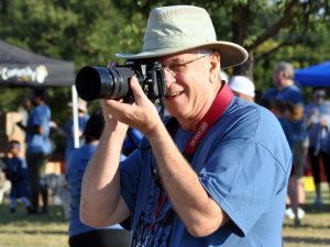 A mature white man wearing a blue t-shirt and tan hat, shoots a photo with a Nikon camera with long lens.