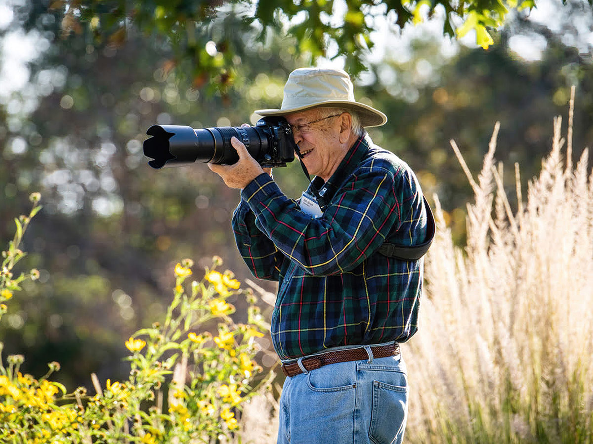 A mature white man wearing a blue plaid shirt, jeans and tan hat, shoots a photo with a Nikon camera with long lens. Behind him are glowing golden grasses and sunflowers.