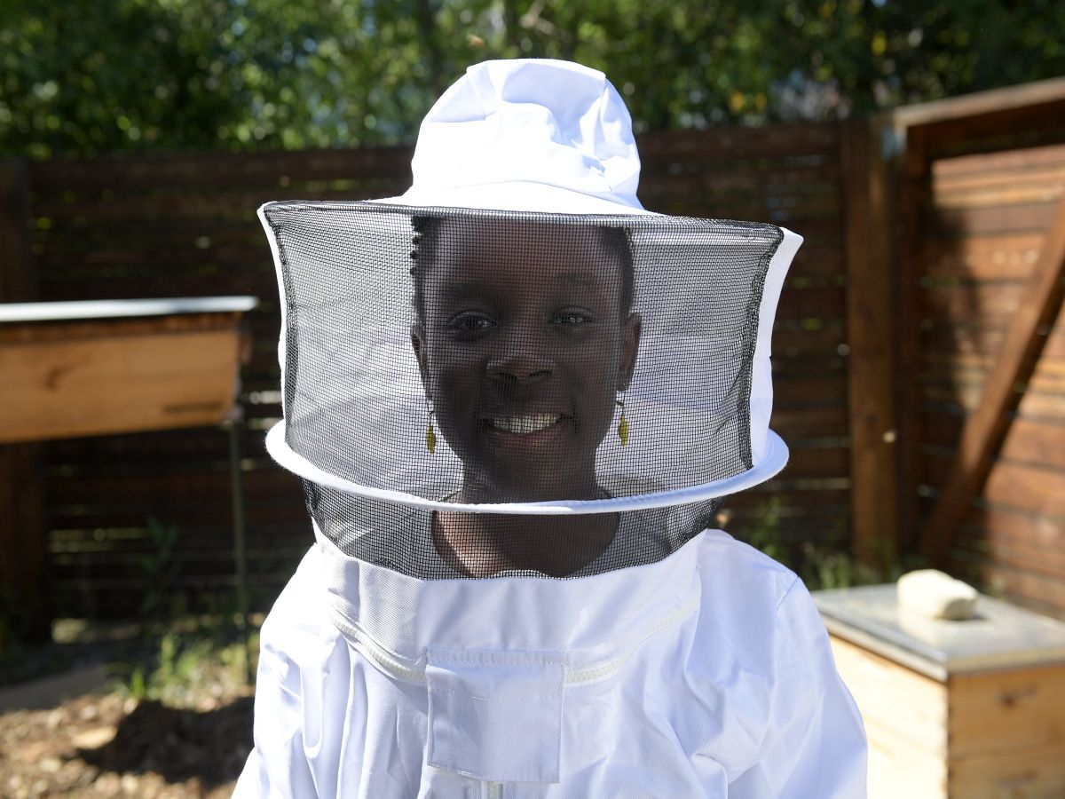 Mikaila Ulmer wearing a white beekeeping suit standing in front of beehives outside