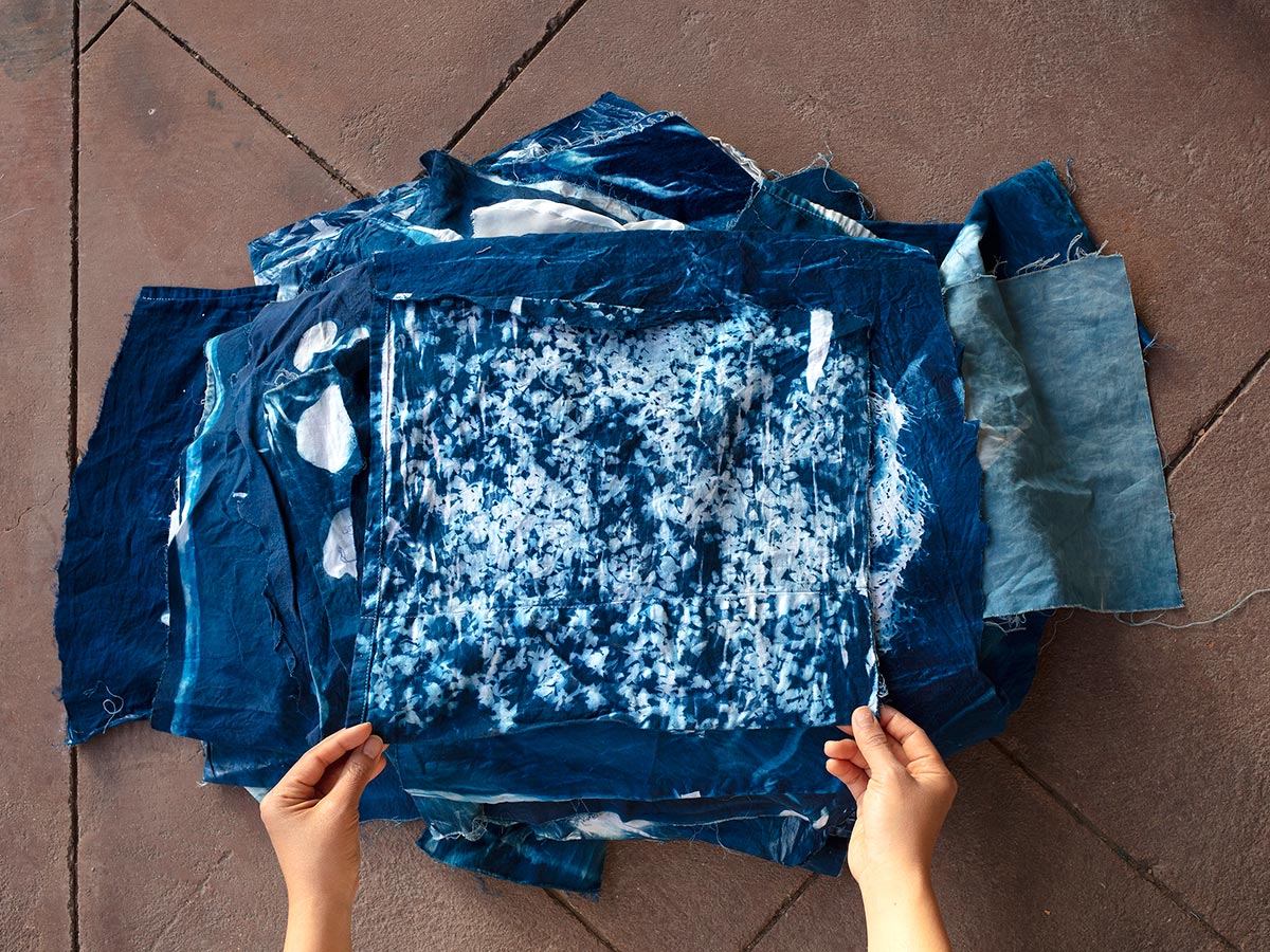 A pile of cyanotypes on fabric with two hands holding the bottom corners of the top piece. Cyanotypes are def blue with white patterns where the plants laid on the fabric.