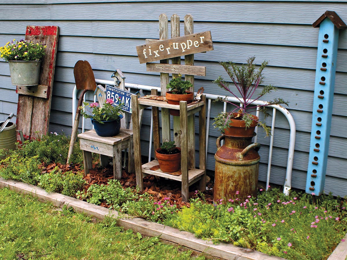 Repurposed items such as rusted watering cans, pieces of worn lumber, an old wooden table and door, a tall and slender birdhouse with many holes, and a sign that says 