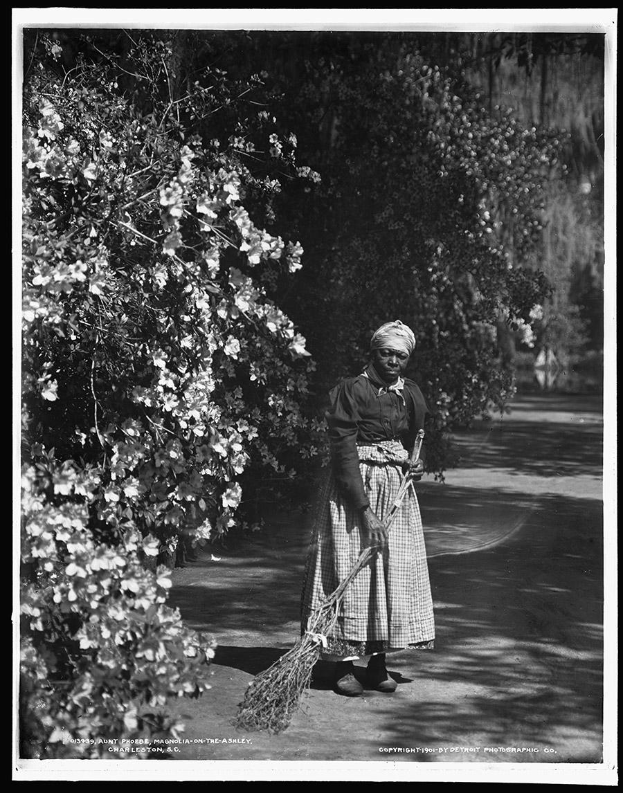 In a black-and-white photo, a Black woman in late-1800s clothing, stands with a broom made of natural materials stands next to a blooming tree.