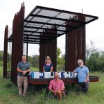 Four staff members stand at an iron structure in a field; the structure has a raised floor lined with binders, showing all the data collected in the area.