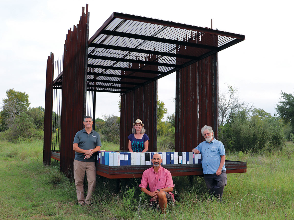 Four staff members stand at an iron structure in a field; the structure has a raised floor lined with binders, showing all the data collected in the area.