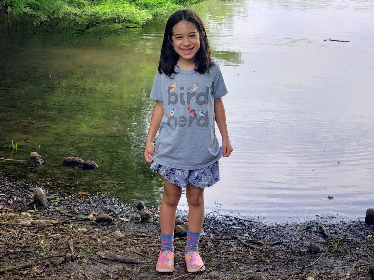 A smiling young girl wearing a "Bird Nerd" shirt stands in front of a riverbed.
