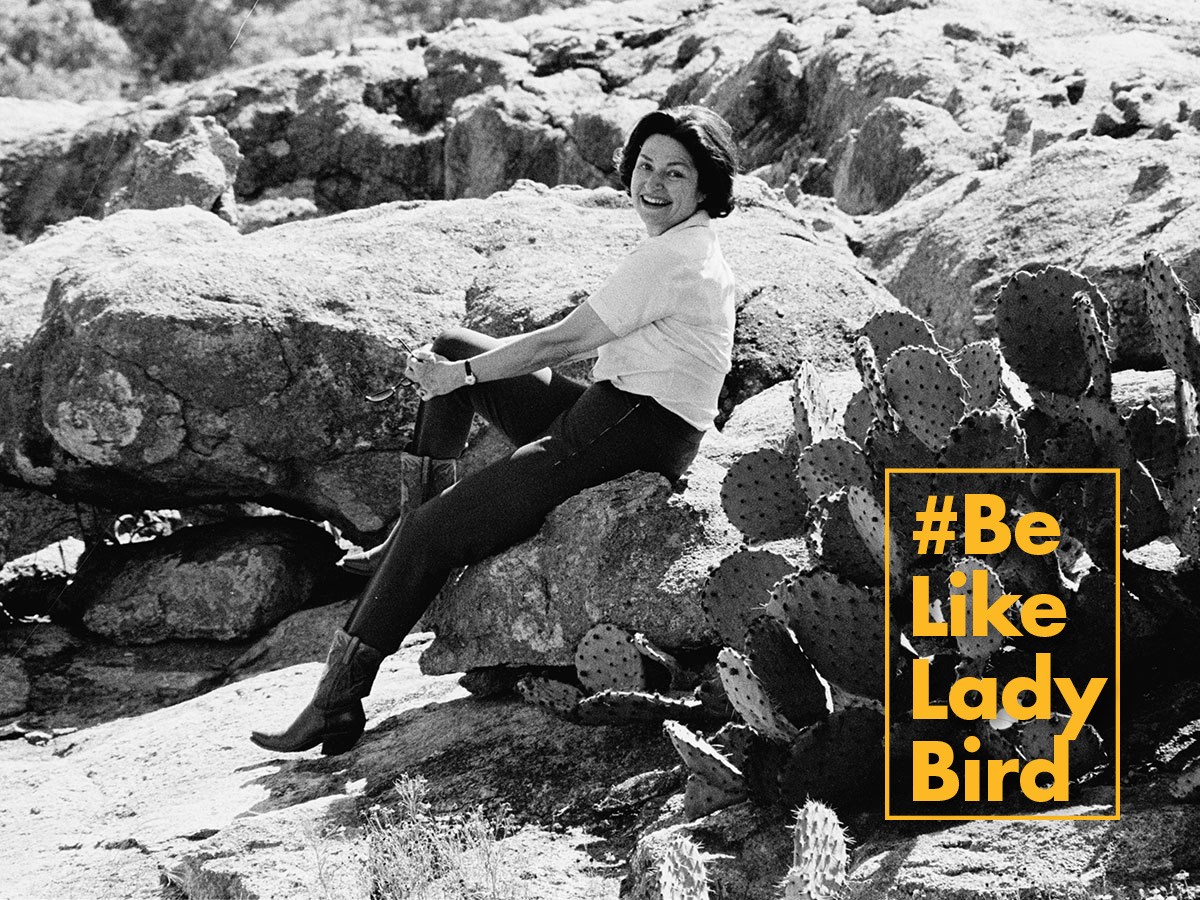 A black and white photo of Lady Bird Johnson sitting on large boulders near prickly pear cacti. #BeLikeLadyBird is in a yellow graphic on the lower right corner.