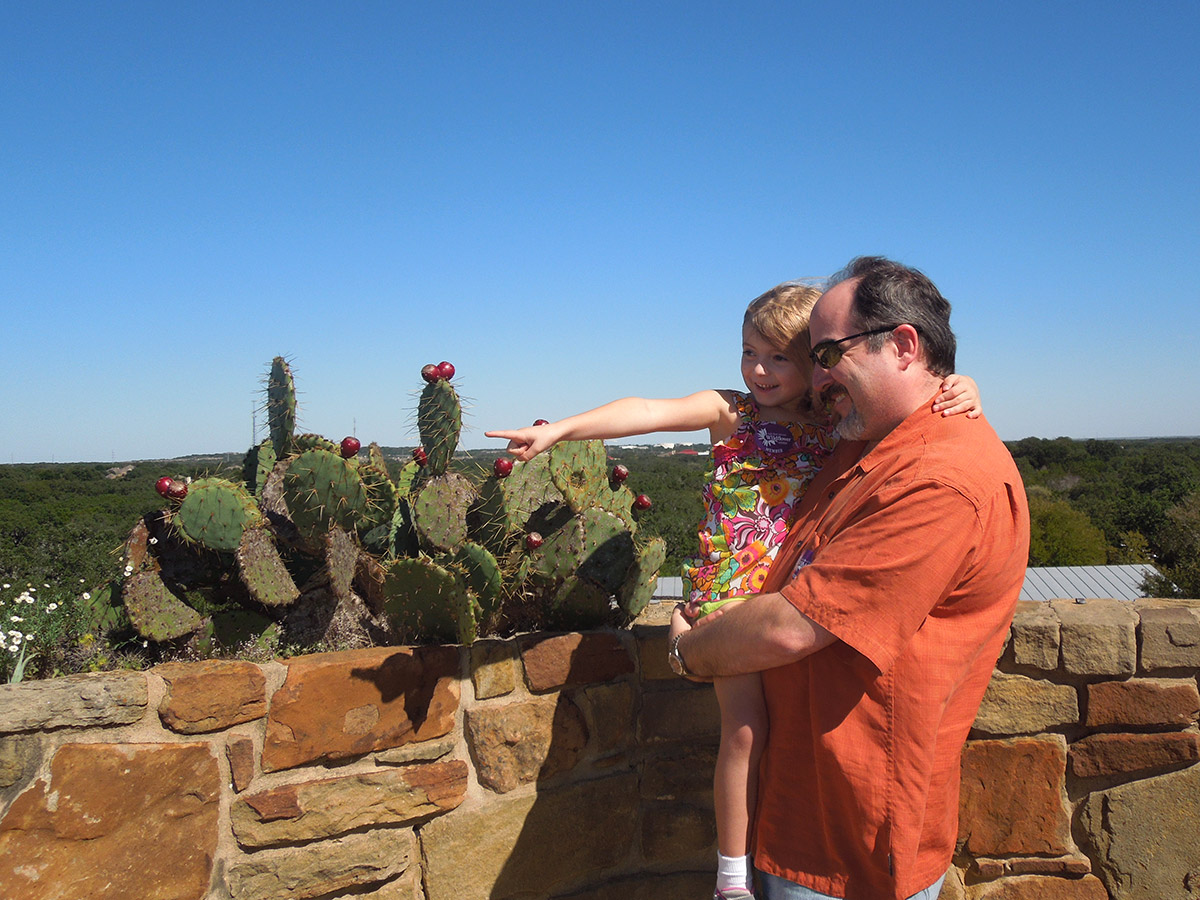A father in an orange shirt holding his young daughter in a flowery dress. They are at the top of our Observation tower and she is pointing towards something out of frame.