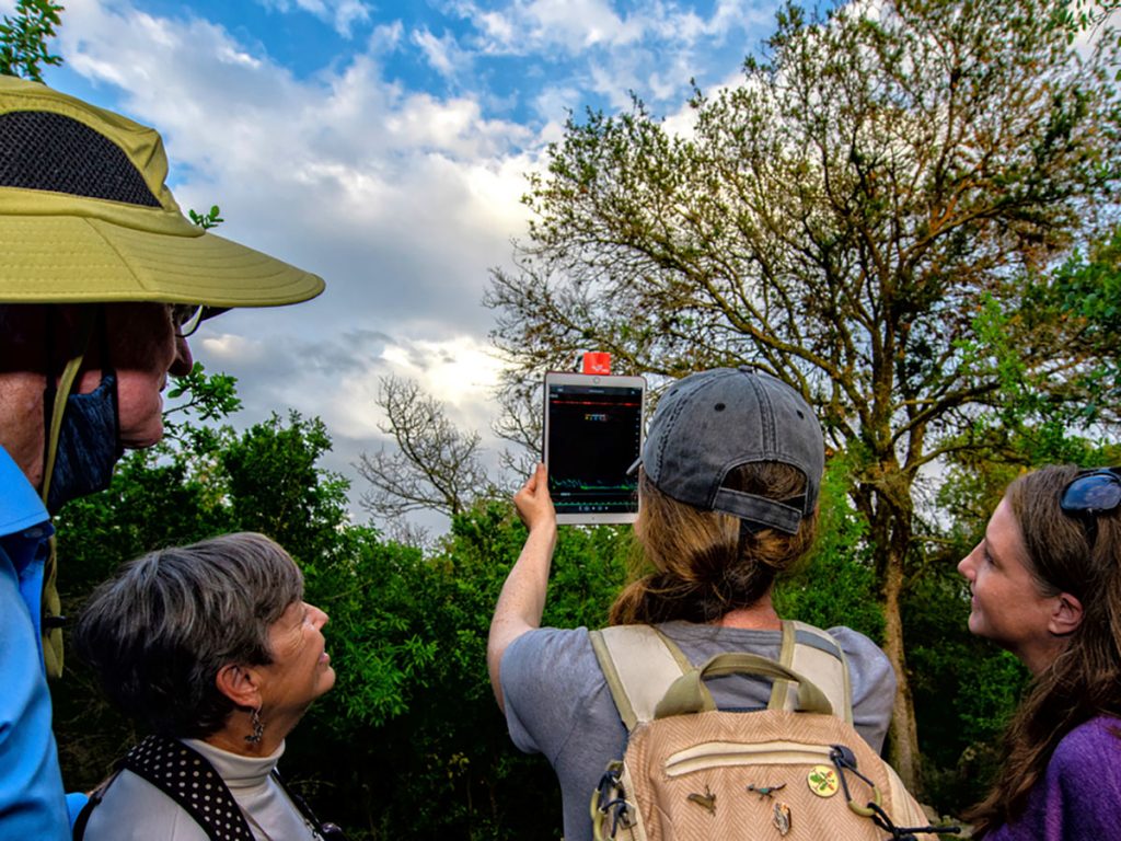 A group of people gathered around an iPad in the middle of a clearing of trees, looking at environmental metrics onscreen.