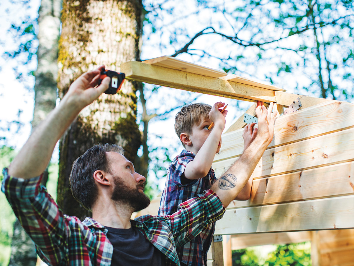 A father and young boy, both wearing paid shirts are measuring a board as they build a treehouse together.