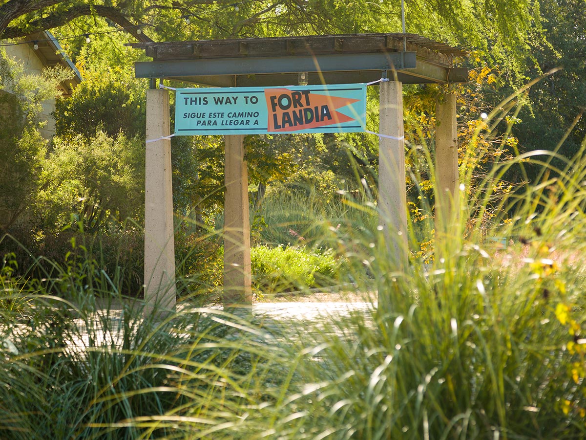 A light blue banner hangs from a pergola surrounded by sunlit grasses and trees, directing visitors to Fortlandia.