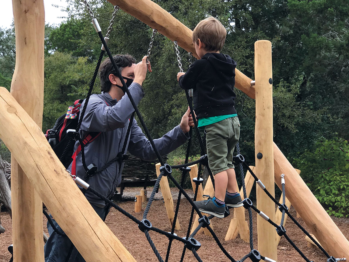 A other and son climbing the black rope webbing secured to the logs of a play structure.