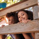 Two young children with dark curly hair peaking between two boards of a treehouse.