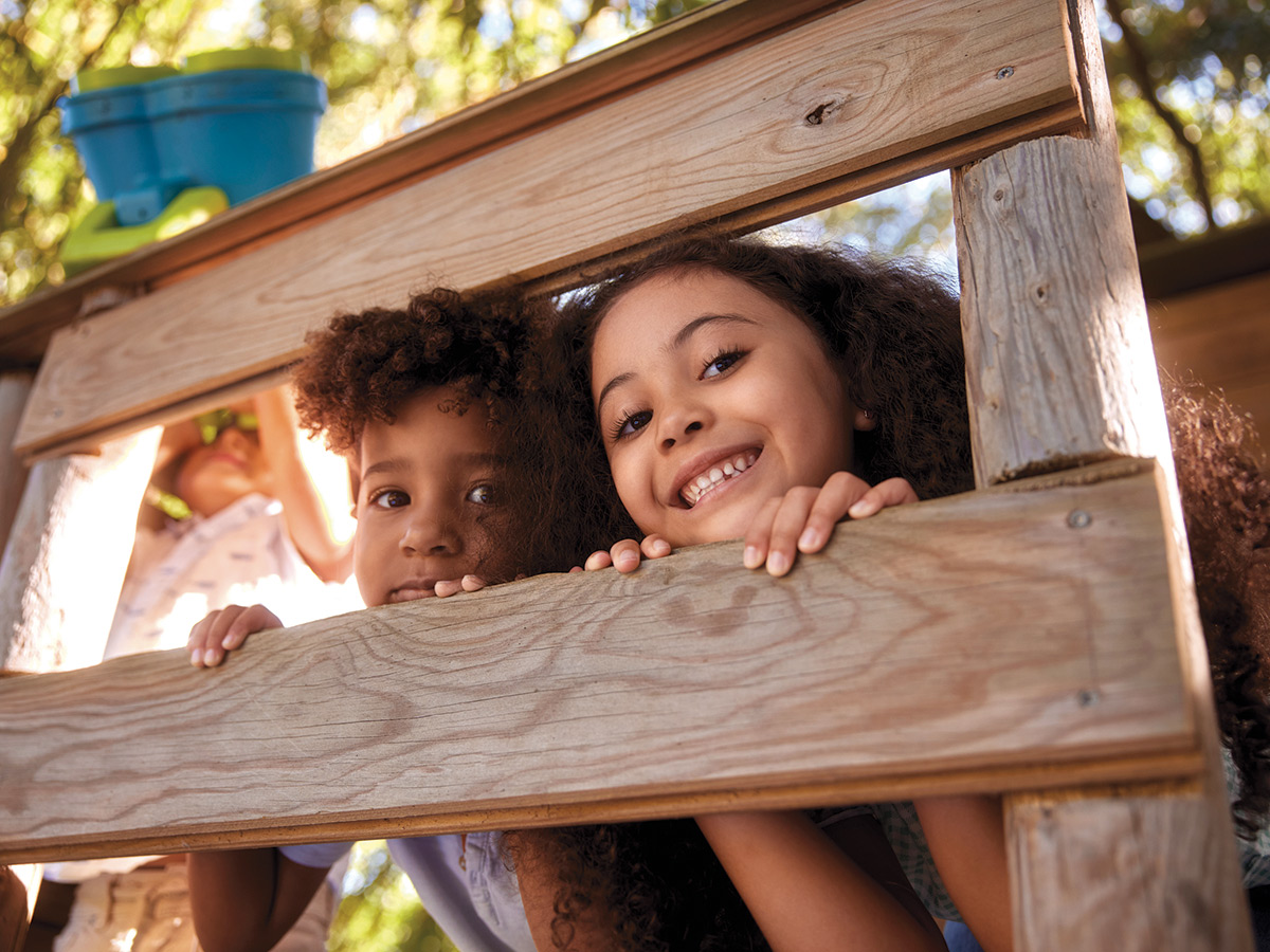 Two young children with dark curly hair peaking between two boards of a treehouse.