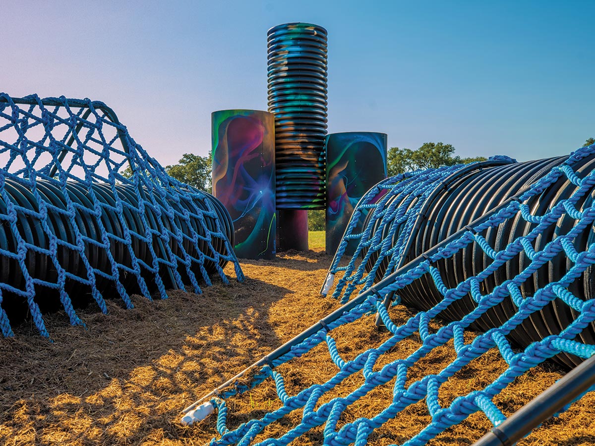 A fort made out of large corrugated, black plastic tubes. They have been spray painted in a graffiti style with bright colors. There is also bright blue rope webbing across some of the tubes for climbing.