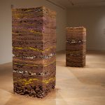A photo of two sculptures free standing in a gallery. They are both vertical columns of clay, soil, stoneware, earthenware and gold in compressed layers. They are mostly a rich brown, but have many layers, a few pink and some gold. Some layers look like broken pieces of pottery.